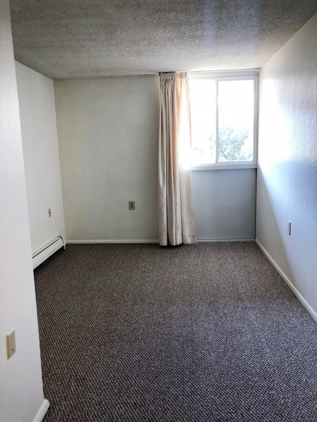 An empty room with a carpeted floor, white walls, and a window with curtains.
