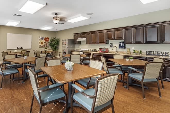 A community dining area with several tables and chairs, featuring a kitchen with wooden cabinets in the background.