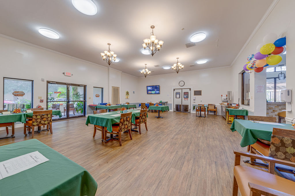 A spacious dining room featuring several tables and chairs, illuminated by chandeliers, with large windows providing natural light.