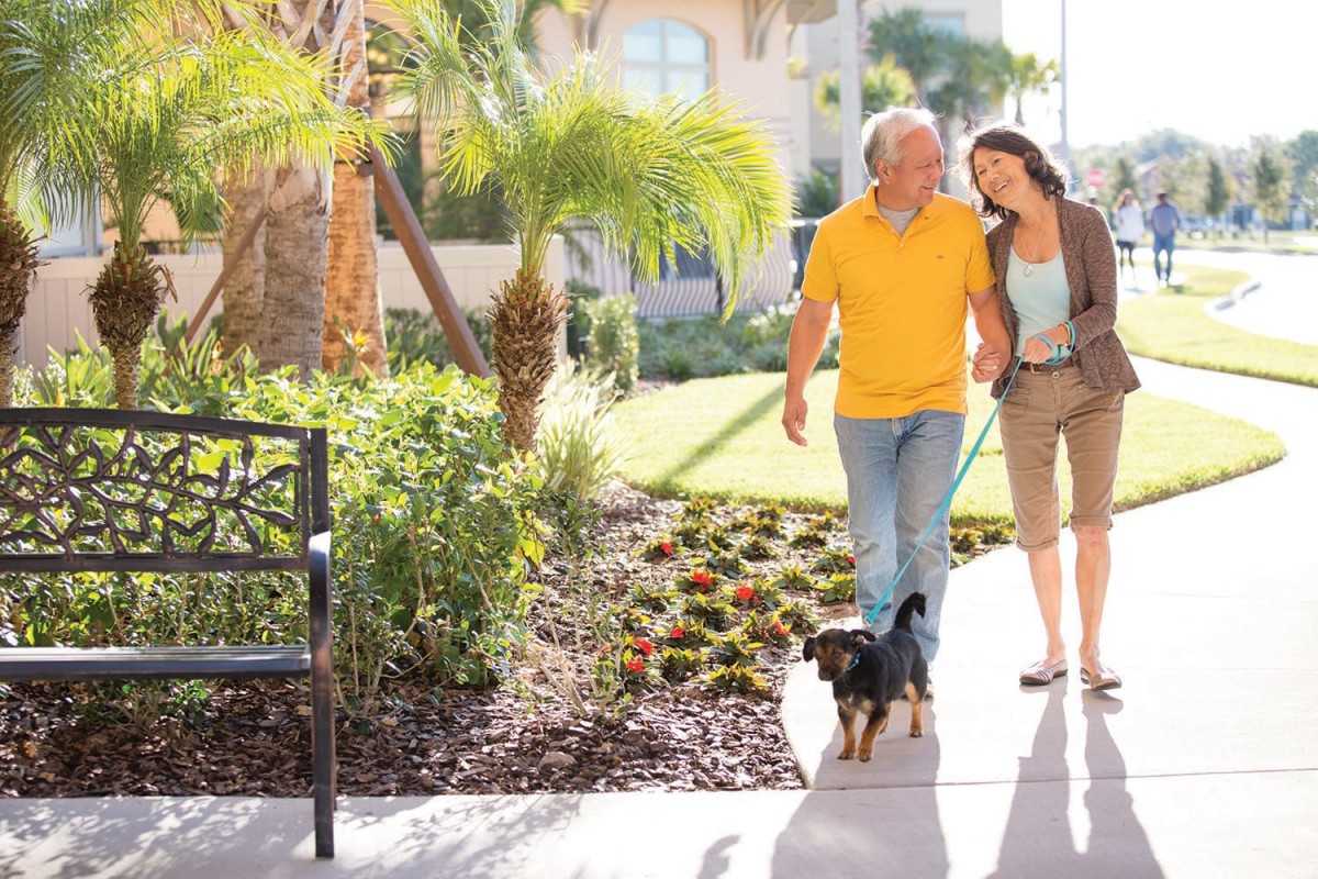 A couple enjoys a sunny day walking their dog in a lush garden setting.