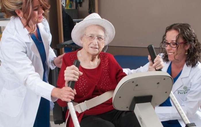 An elderly woman receiving assistance while exercising on gym equipment.