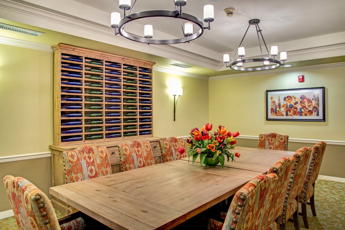 A warmly lit dining room with a large wooden table, surrounded by patterned chairs and adorned with chandeliers and a floral centerpiece.