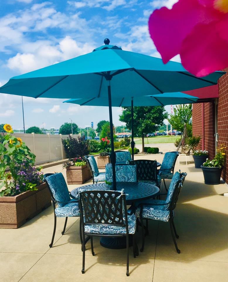 A sunny patio with outdoor seating and a blue umbrella, surrounded by potted plants.