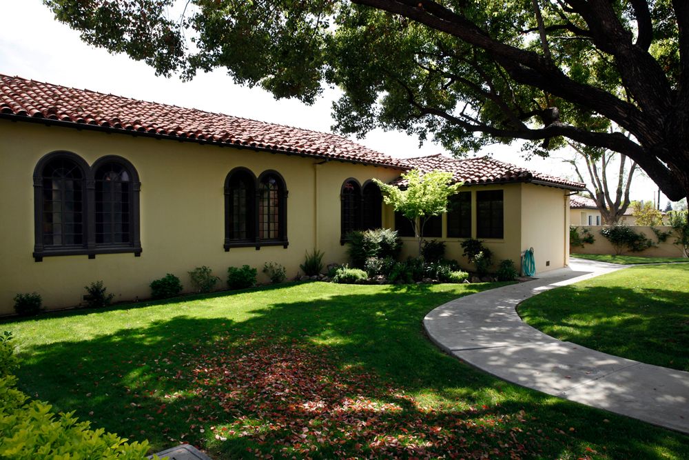 A charming house with a red-tiled roof surrounded by a lush garden and shaded by large trees.