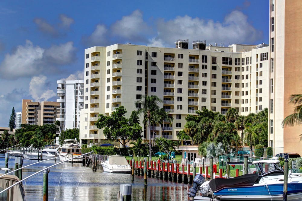 A scenic waterfront view featuring a residential apartment building with boats docked nearby and palm trees lining the shore.