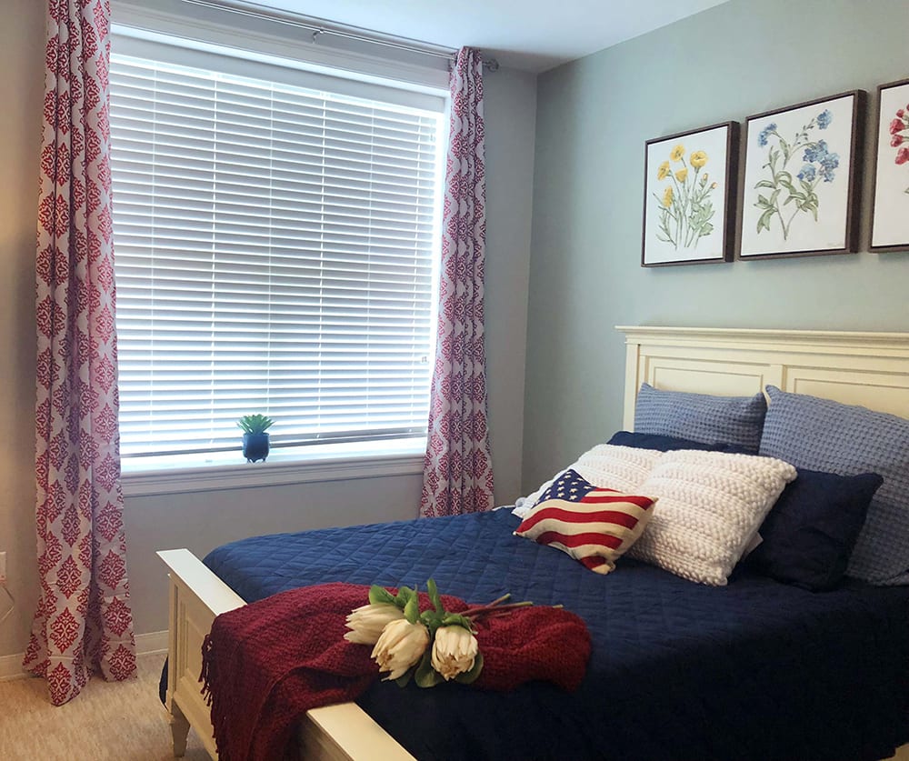 A cozy bedroom with a patriotic-themed bedspread and decor, featuring a large window with drawn curtains and elegant artwork above the bed.