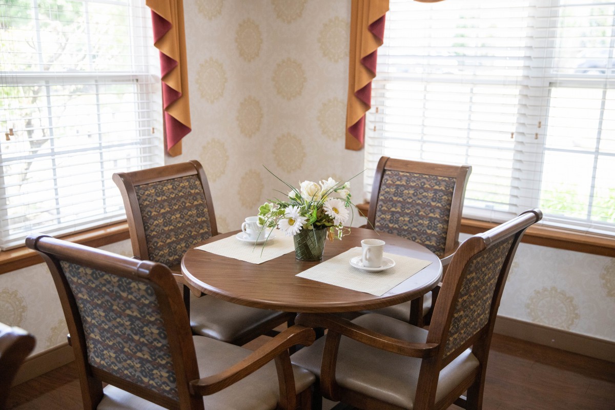 A cozy dining area with a wooden table, four chairs, and a floral centerpiece by a window with curtains.