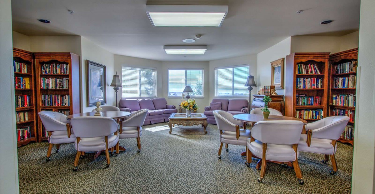 A cozy living room with sofas and tables arranged around a central area, featuring large windows and soft lighting.