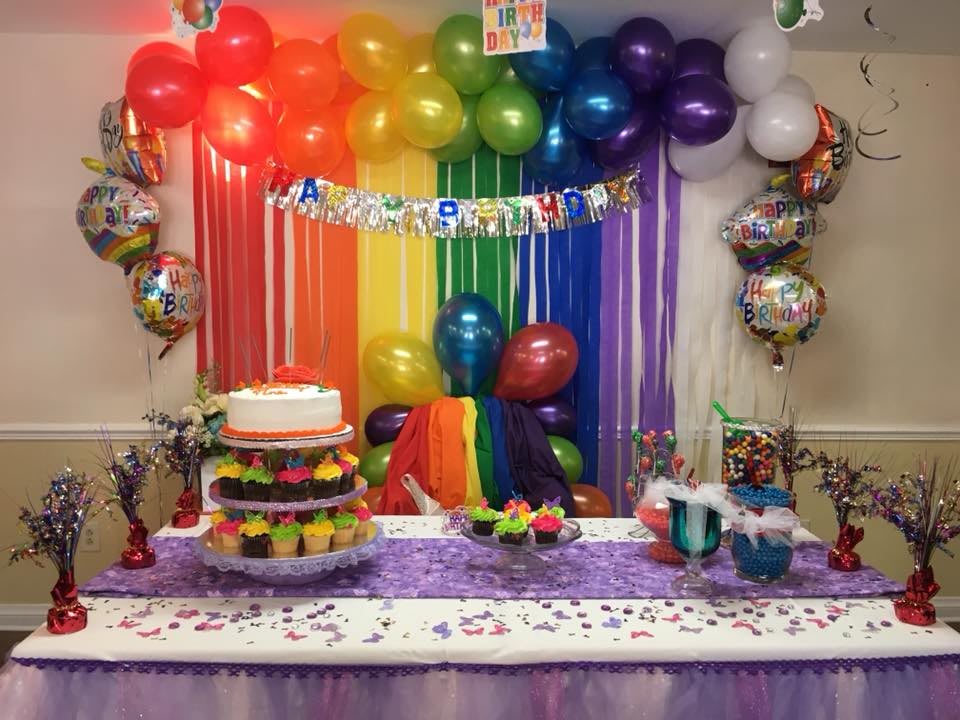 A festive party setup with a rainbow theme featuring colorful balloons, streamers, and a decorated cake.