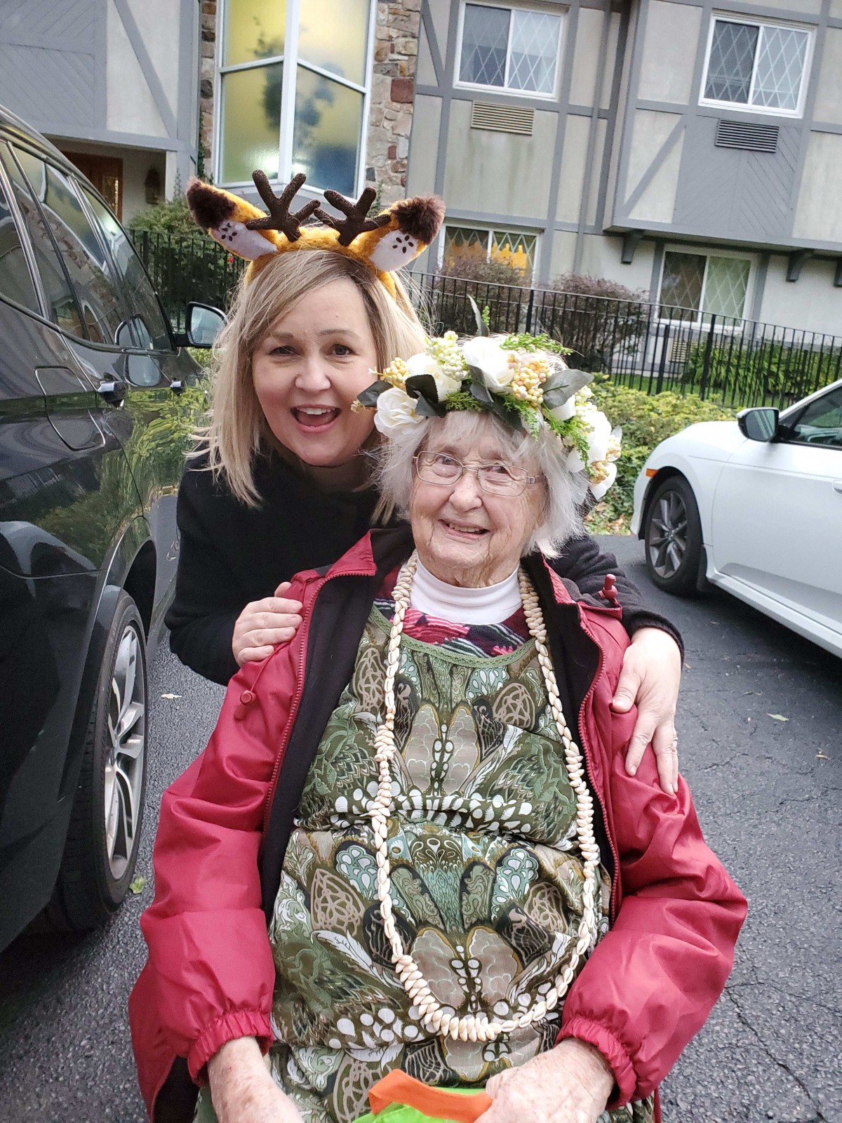 An elderly woman with a flower crown sits in a wheelchair, accompanied by a smiling younger woman wearing a giraffe headband.