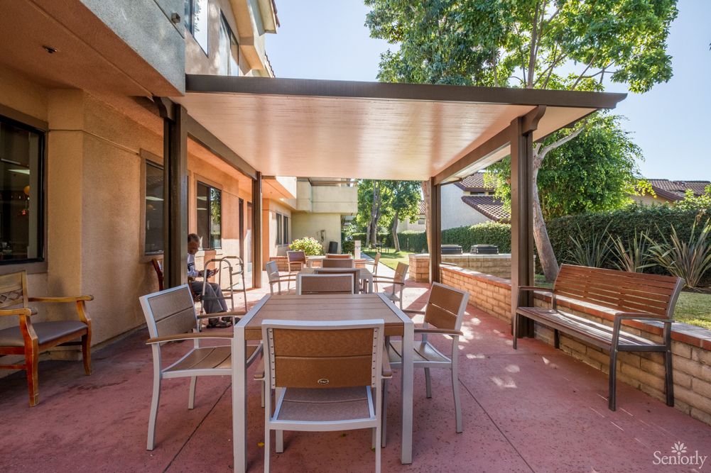 A sunny outdoor patio with tables and chairs under a pergola, surrounded by trees.