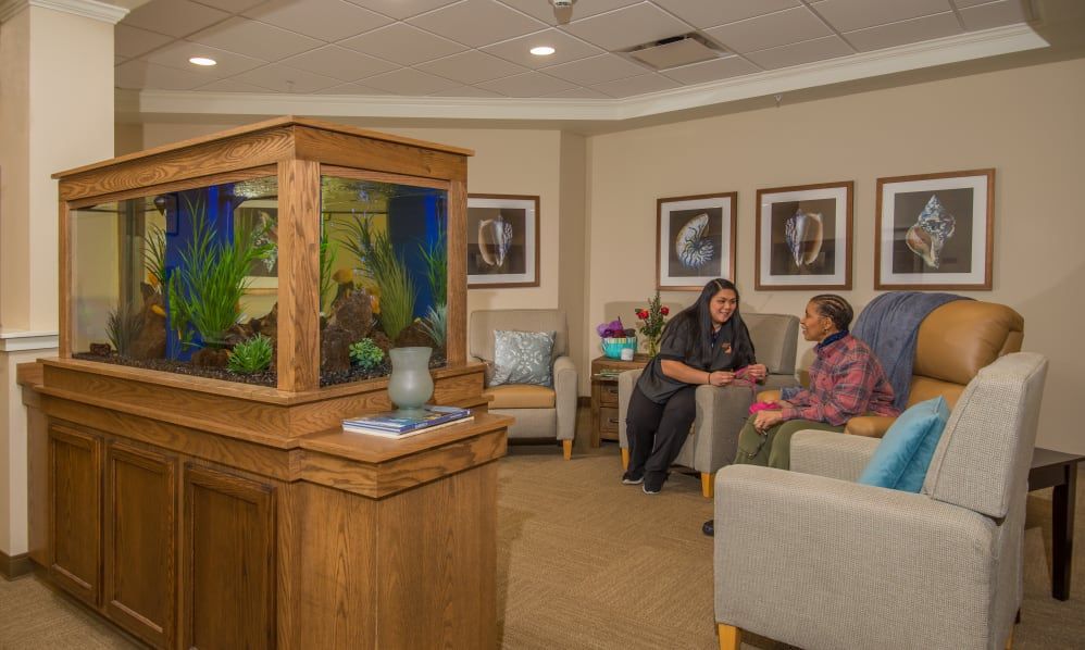 Two people converse in a cozy waiting room with an aquarium centerpiece and comfortable seating.
