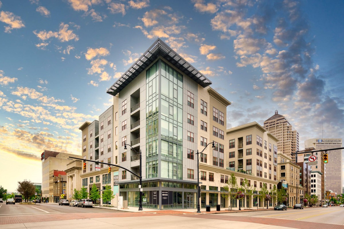 A modern urban apartment building with a glass facade, set against a backdrop of a partly cloudy sky.