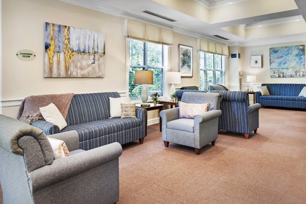 A cozy living room with comfortable sofas and armchairs, featuring a carpeted floor and large windows accented by elegant decor.