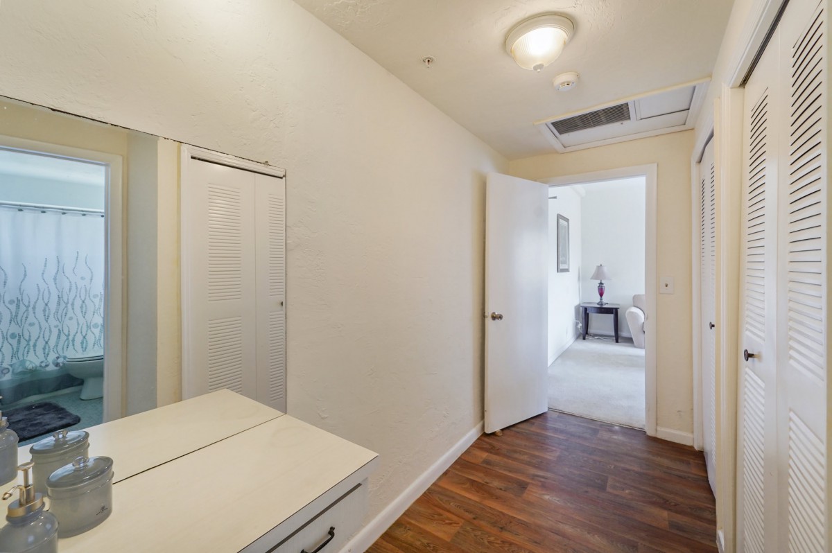 A hallway featuring a wooden floor, a mirror, and a light fixture on the ceiling. An open door leads to another room.