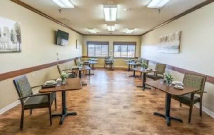 A clean and well-lit dining room with several tables and chairs, featuring large windows and a wooden floor.