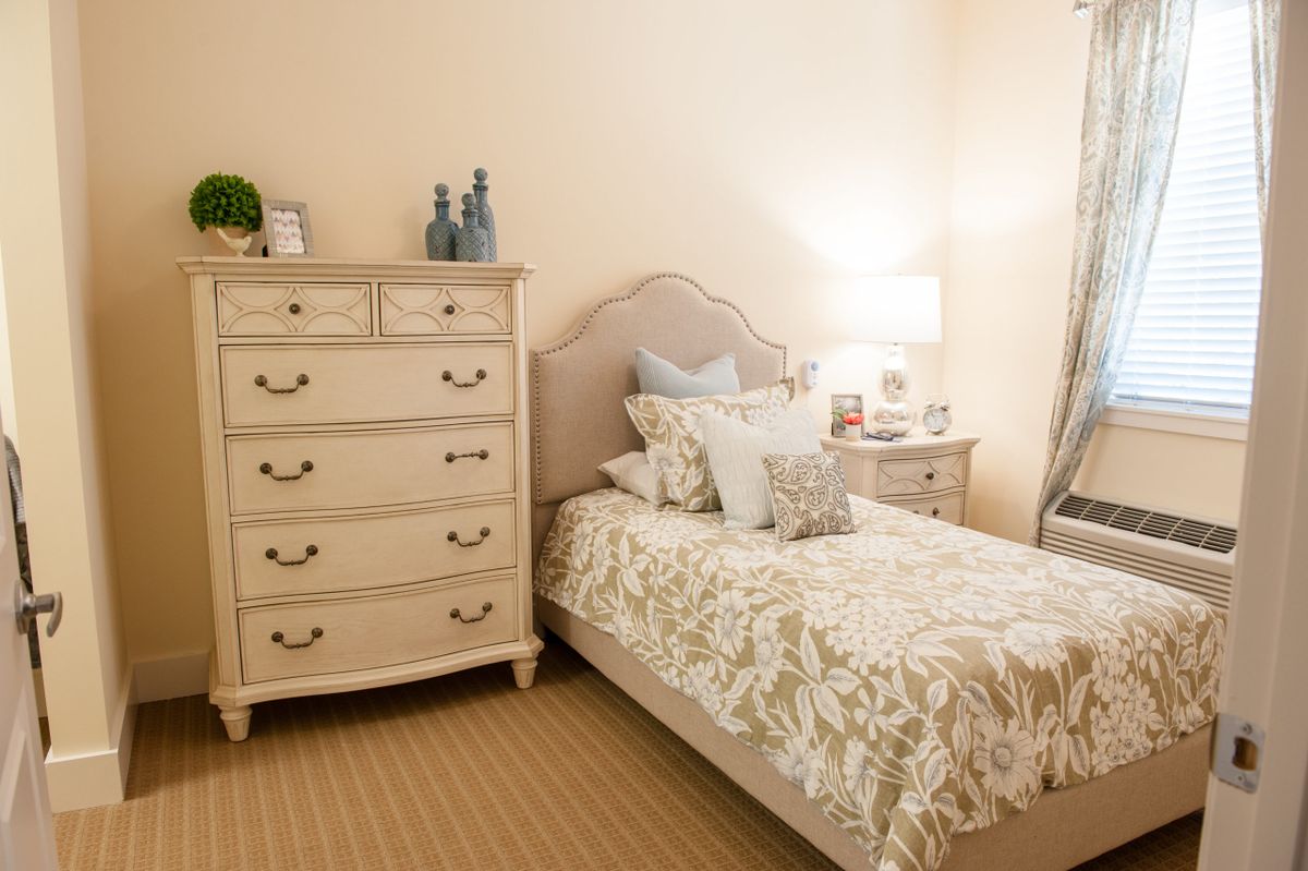 A cozy bedroom with a single bed, decorative pillows, a dresser, and a lamp.