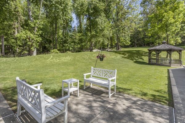 A peaceful garden patio with white outdoor benches, surrounded by lush greenery on a sunny day.