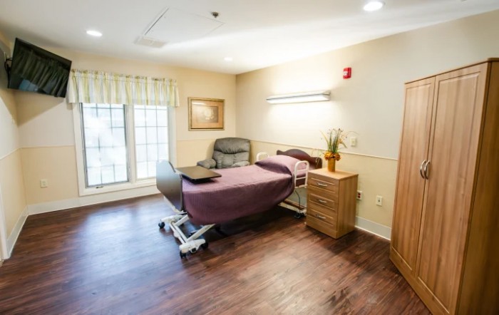 A clean and well-lit hospital room with a bed, chair, and wooden flooring.