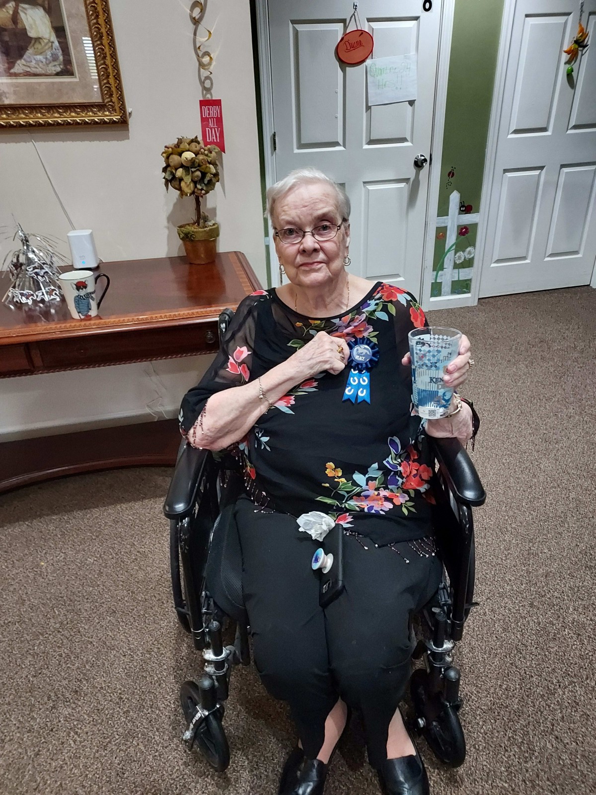 An elderly woman seated in a wheelchair displays an award ribbon while smiling indoors.