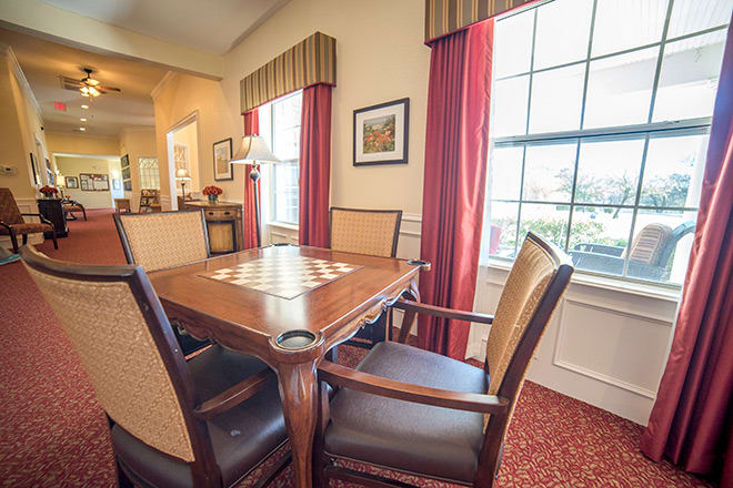 A cozy dining room with a wooden table and upholstered chairs, featuring large windows with red curtains.