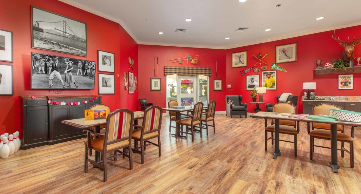 A cozy dining area with red walls, decorated with framed pictures and featuring multiple tables and chairs on a wooden floor.