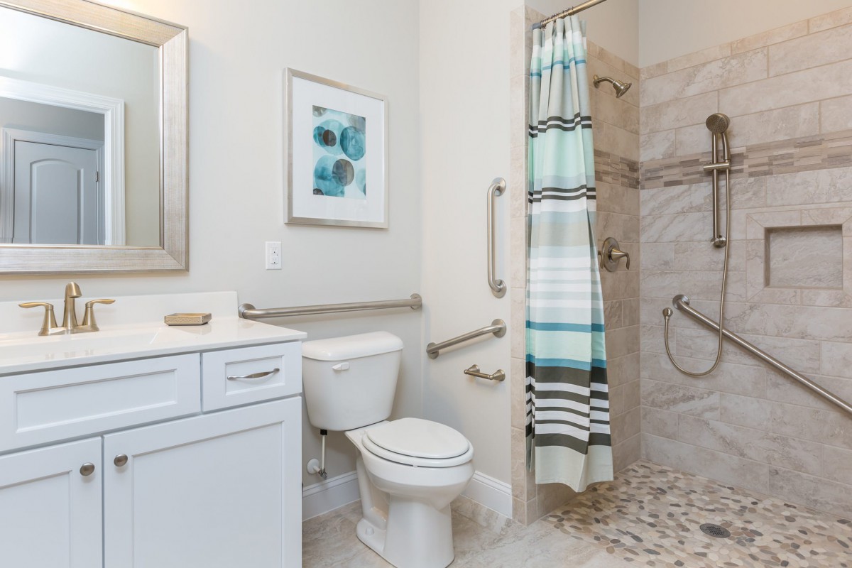 A modern bathroom featuring a shower with grab bars, a toilet, and a sink with storage.