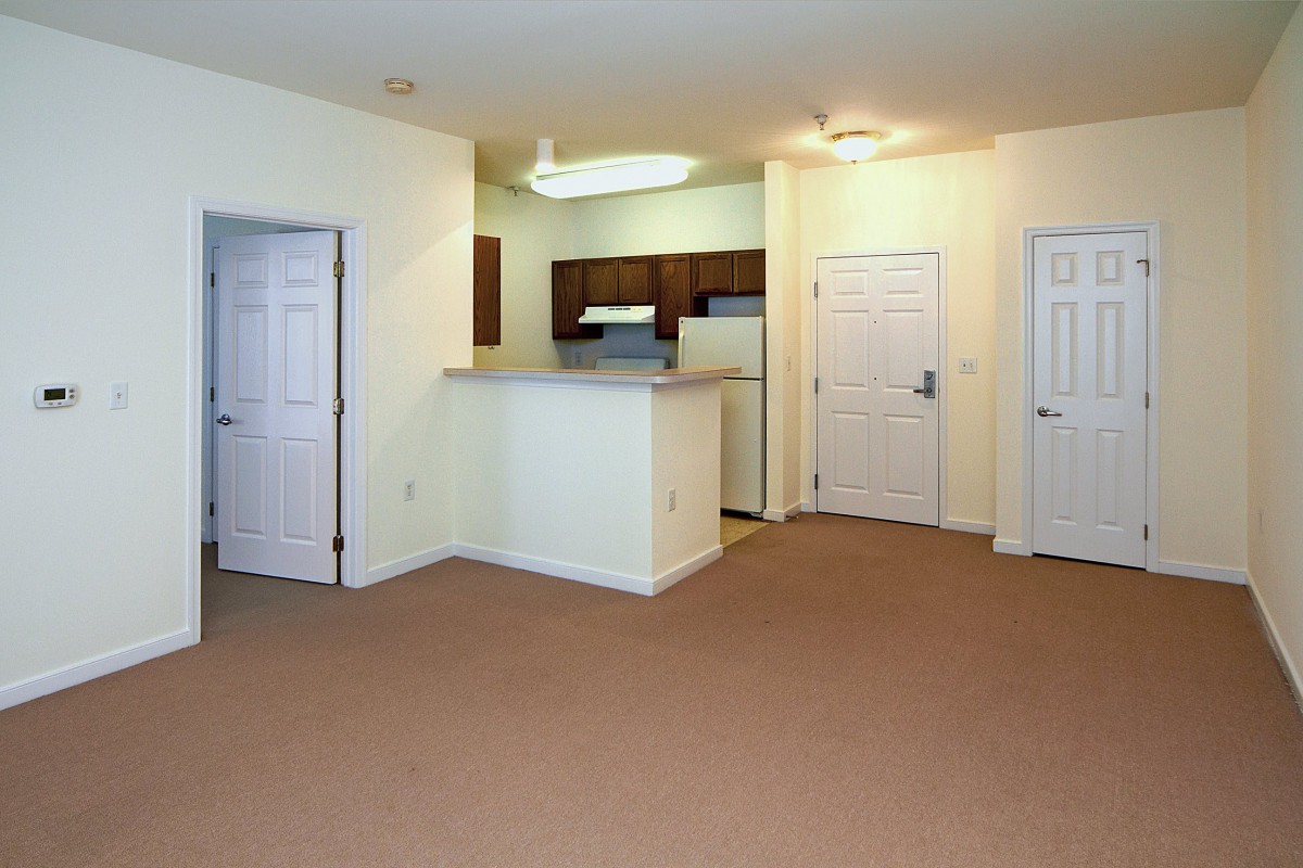 An unfurnished apartment with a small kitchen area, white walls, and carpeted flooring.