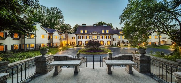A grand mansion with illuminated exterior and central fountain, surrounded by trees during the evening.