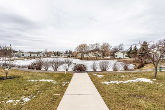 A snowy suburban landscape with a sidewalk leading to a pond, surrounded by houses and bare trees under an overcast sky.