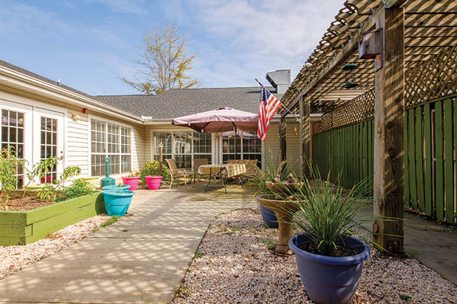 A cozy backyard patio featuring colorful potted plants, outdoor seating under a pergola, and a flag waving in the breeze on a sunny day.