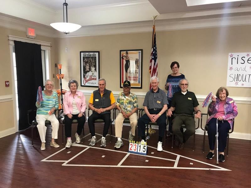 A group of seniors seated together for a community event indoors, with the USA flag in the background.