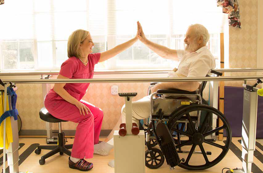 A physical therapist and a senior man in a wheelchair share a high-five during a rehabilitation session.