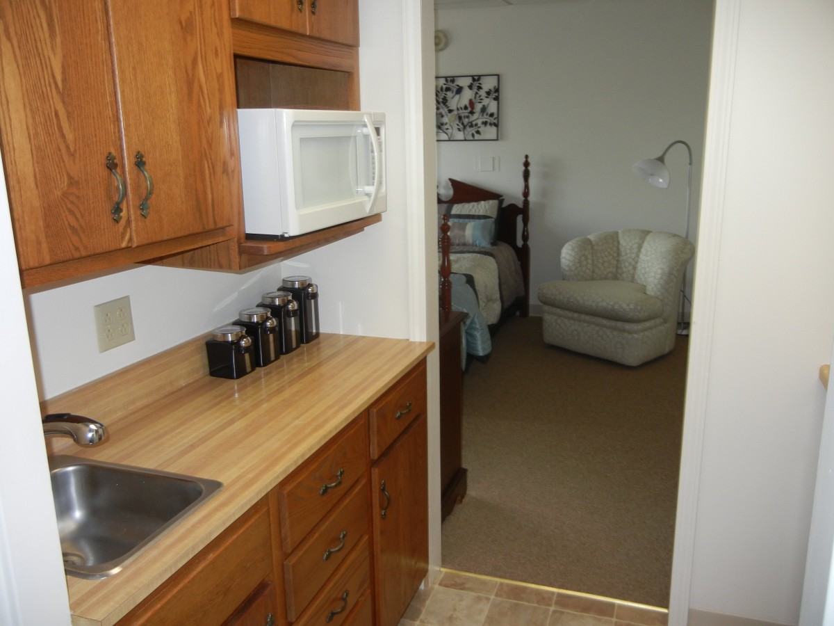 A small kitchen area leading to a cozy living space with a chair and lamp.