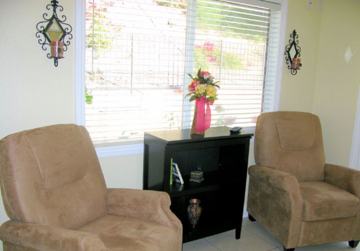 A cozy living room with two armchairs, a cabinet with a flower arrangement in a vase, and natural light streaming through the window blinds.