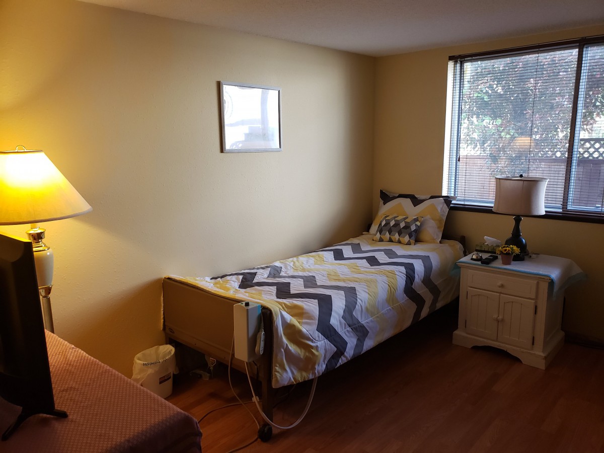 A cozy bedroom featuring a single bed with a chevron-patterned blanket, a nightstand with a lamp, and a window letting in natural light.