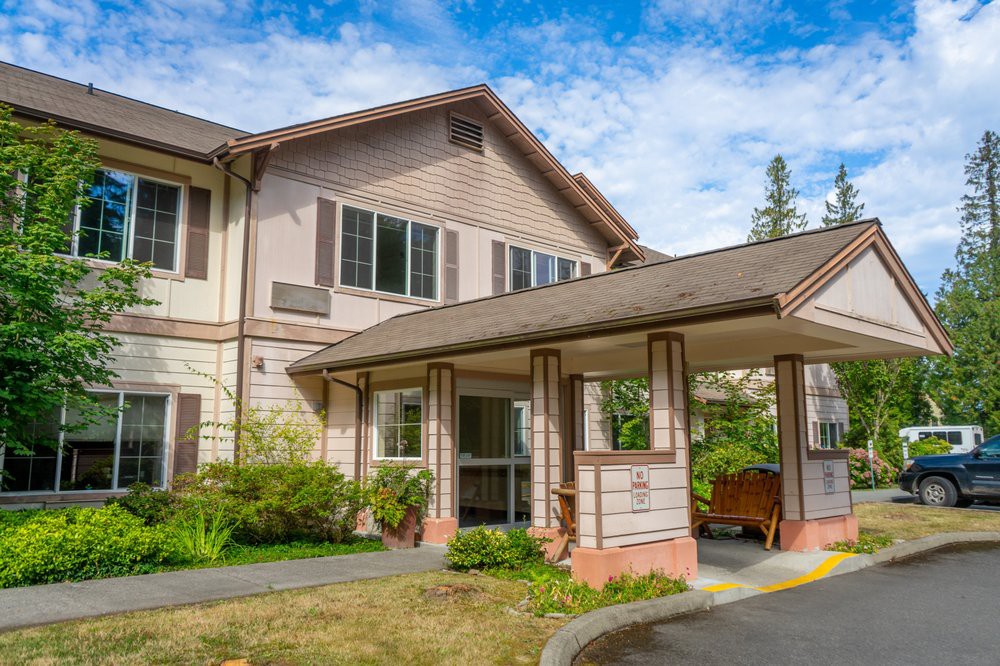 A two-story residential building with a sheltered entrance, surrounded by greenery under a partly cloudy sky.