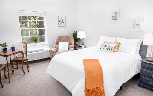 A bright and cozy bedroom featuring white bedding, a comfortable armchair, and natural light streaming in from the window.