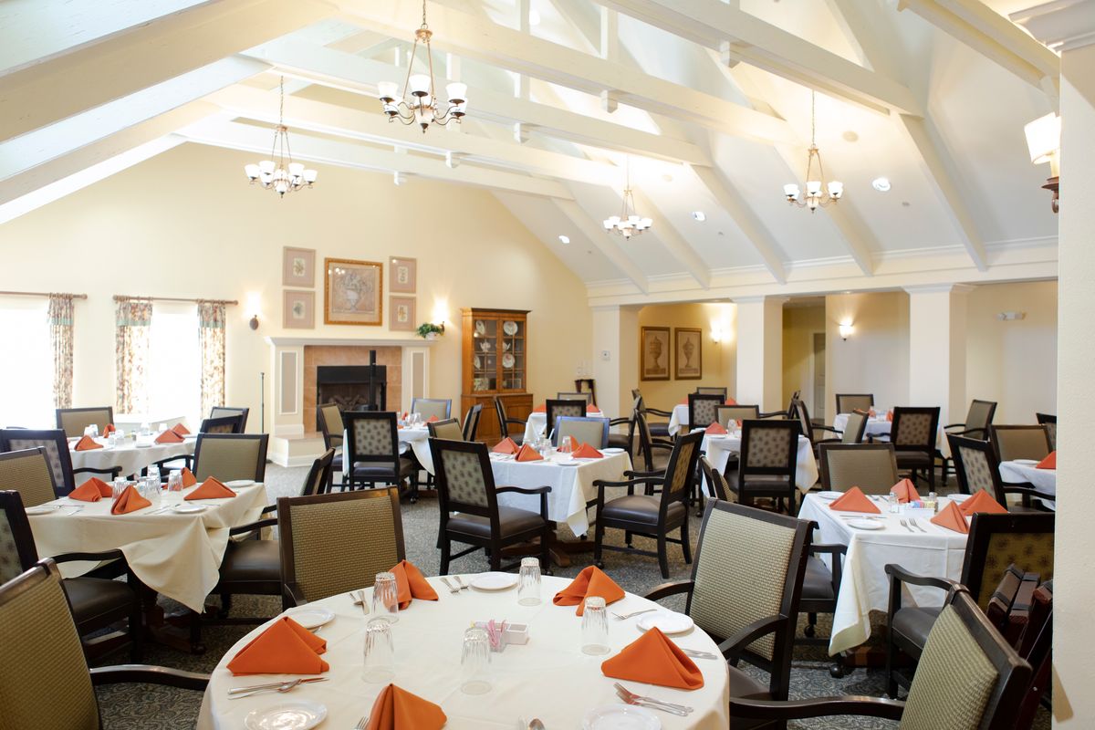 A well-lit dining room with neatly arranged round tables, each set with orange napkins and surrounded by elegant chairs.