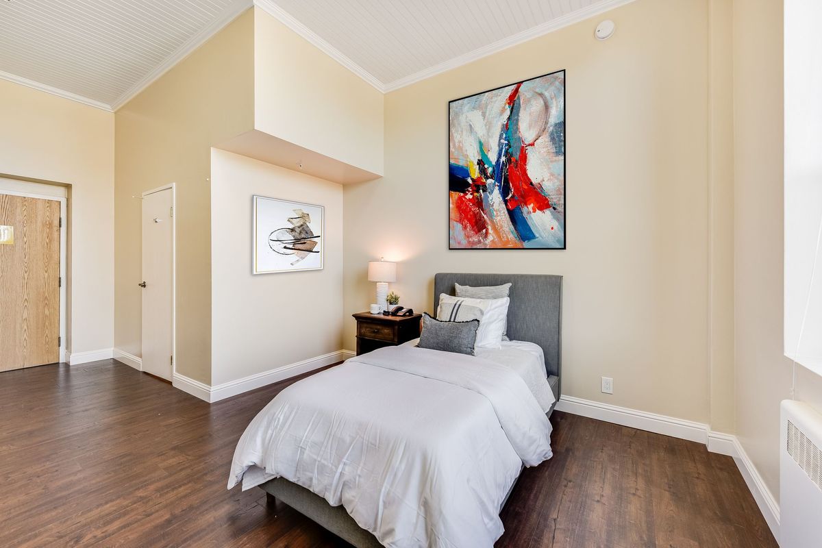 A modern bedroom featuring a single bed with white bedding, complemented by abstract artwork on the walls and a nightstand with a lamp on a hardwood floor.