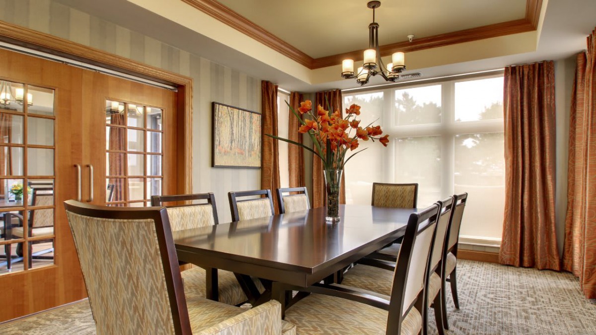 A spacious dining room featuring a large table surrounded by chairs, with elegant lighting from a chandelier and bright natural light streaming in through the windows.