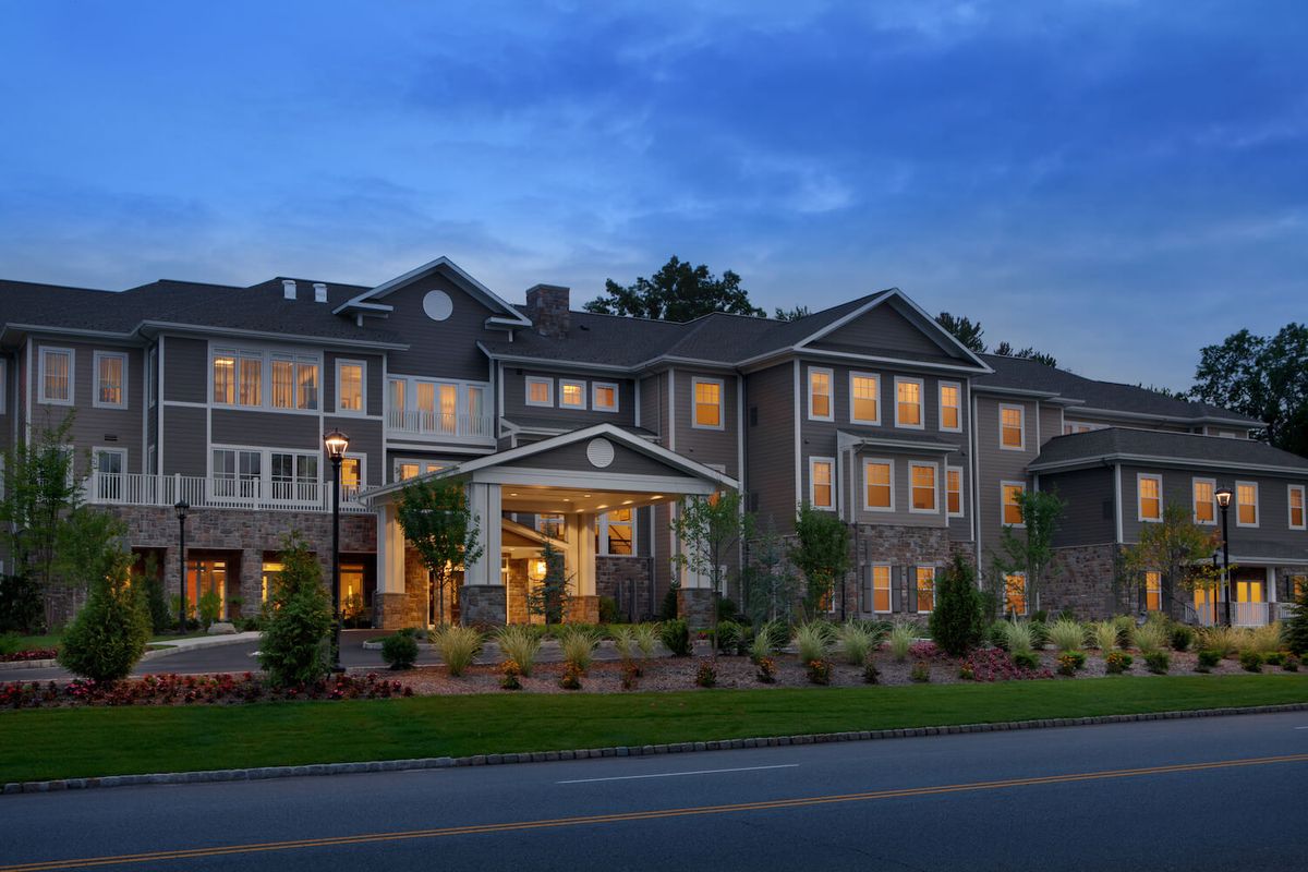 A well-lit residential building in the evening with landscaped surroundings and a road in the foreground.
