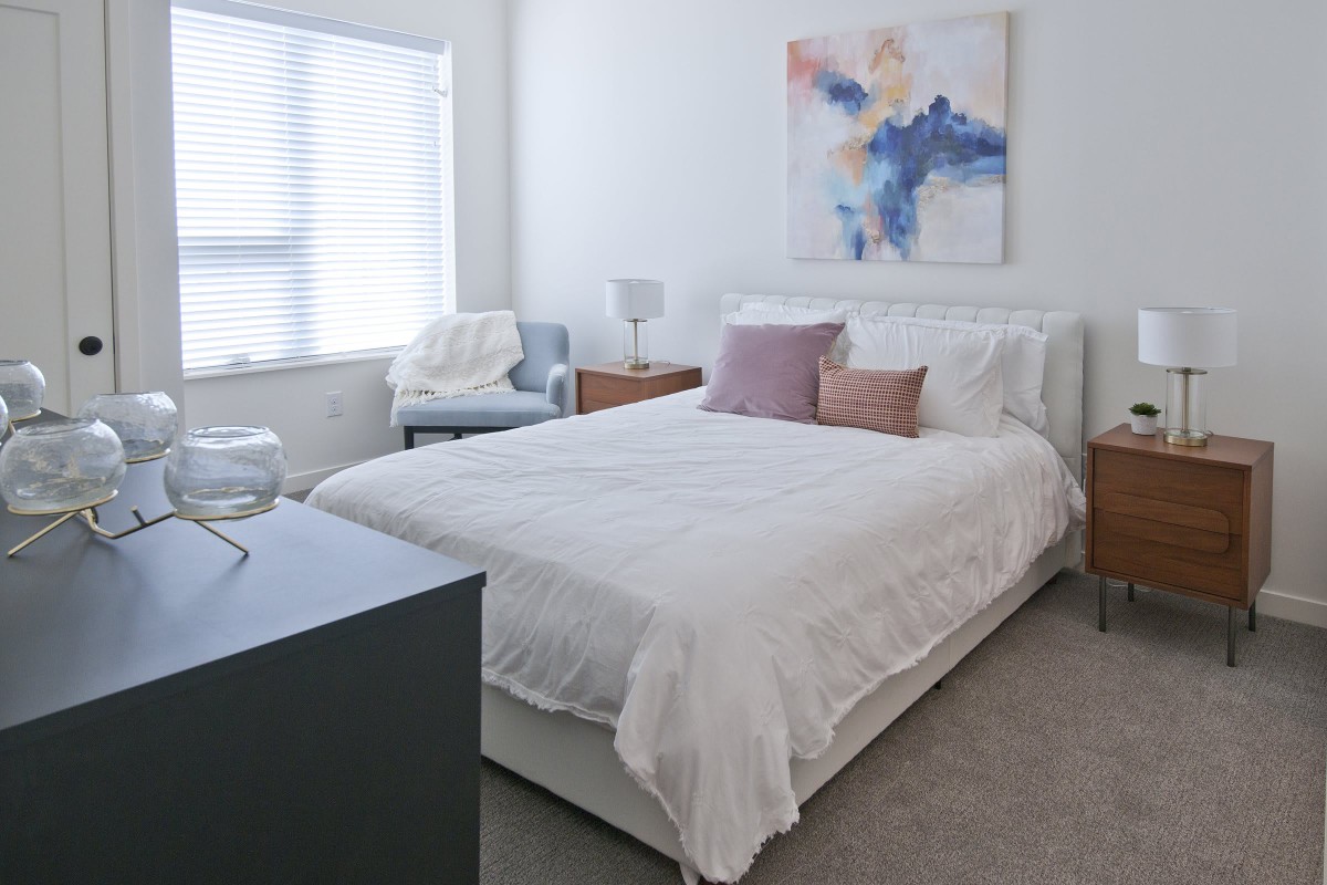 A modern and cozy bedroom with minimalist decor, featuring a neatly made bed, a piece of colorful abstract artwork on the wall, and a comfortable chair by the window.