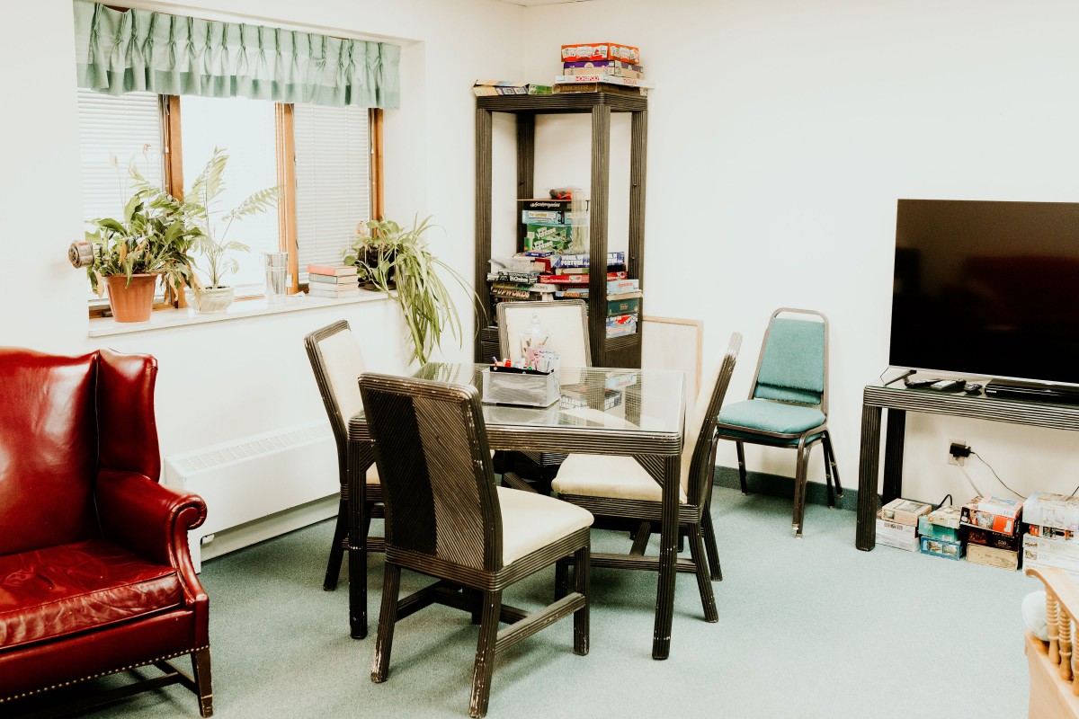 A cozy dining room with a wooden table and chairs, plants on the windowsill, and a tall shelf in the corner.