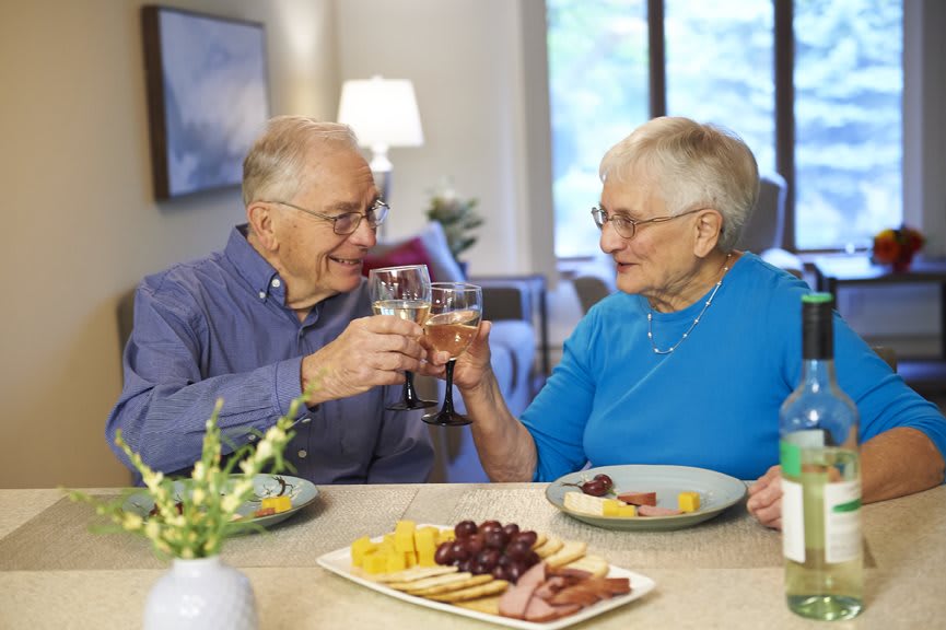 An elderly couple enjoying a meal with a cheese platter, toasting to each other with smiles.