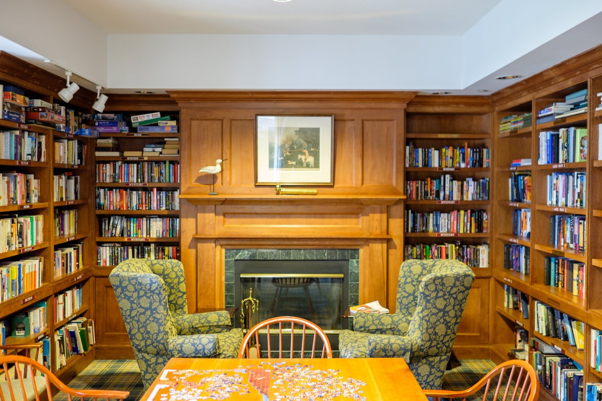 A cozy library room featuring a wooden bookshelf and a fireplace, flanked by two patterned armchairs.