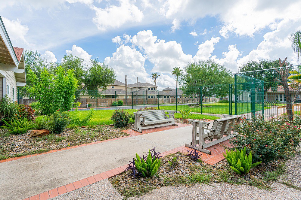 A restful park scene with benches along a pathway, surrounded by trees and greenery under a cloudy sky.