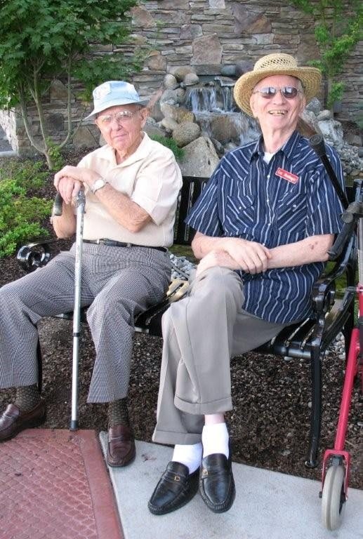 Two elderly men sitting on a bench outdoors, both wearing hats and holding walking canes.