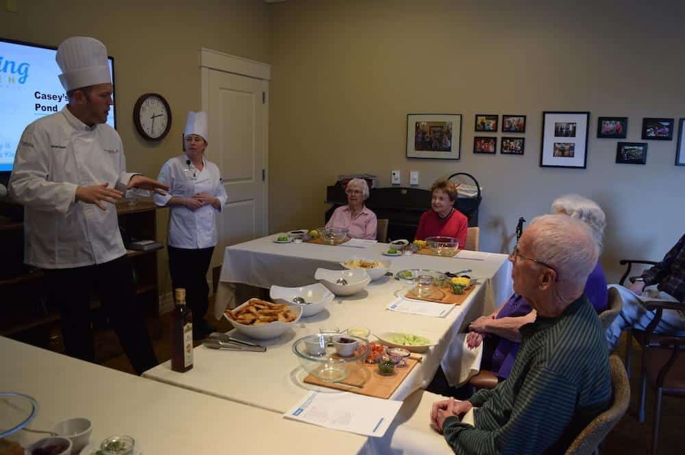 A group of elderly people attending a cooking class with chefs in a cozy room.