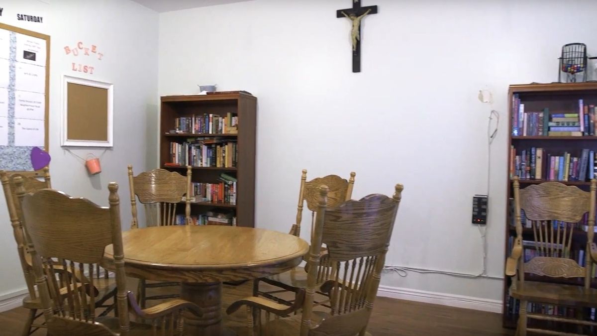 A dining room featuring a round wooden table with chairs, a bookshelf in the corner, and a crucifix on the wall.